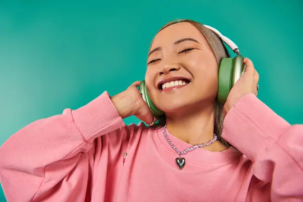 Complacido joven asiático chica en inalámbrico auriculares escuchar música en turquesa fondo - foto de stock