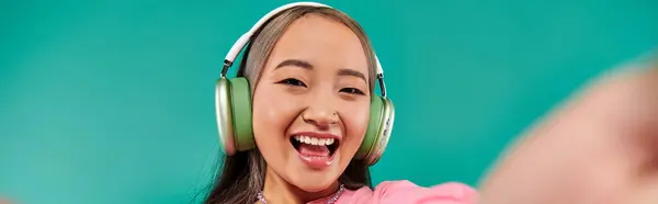 Alegre joven asiático chica en inalámbrico auriculares escuchar música y tomar selfie en turquesa, bandera - foto de stock