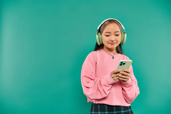 Happy asian girl in wireless headphones listening music and using smartphone on turquoise backdrop — Stock Photo