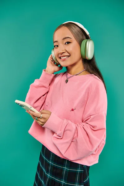 Chica asiática positiva en auriculares inalámbricos escuchar música y el uso de teléfono inteligente en turquesa - foto de stock