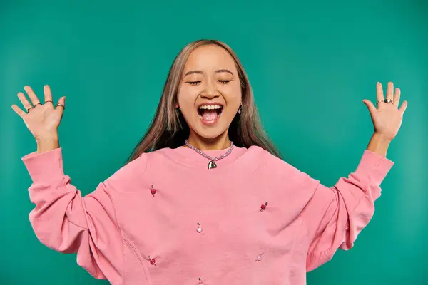 Portrait of excited and young asian girl in pink sweatshirt posing on turquoise background — Stock Photo