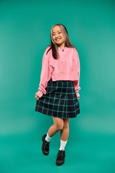 Longitud completa de feliz y joven chica asiática en sudadera rosa posando sobre fondo turquesa - foto de stock