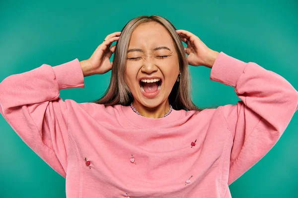 Retrato de emocional joven asiático chica en rosa sudadera gritando en turquesa fondo - foto de stock