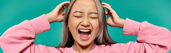Portrait of emotional young asian girl in pink sweatshirt screaming on turquoise background, banner — Stock Photo