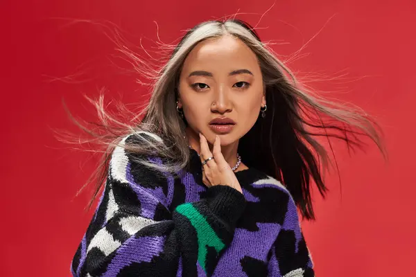 Charming young asian woman in vibrant sweater with animal print touching cheek on red backdrop — Stock Photo
