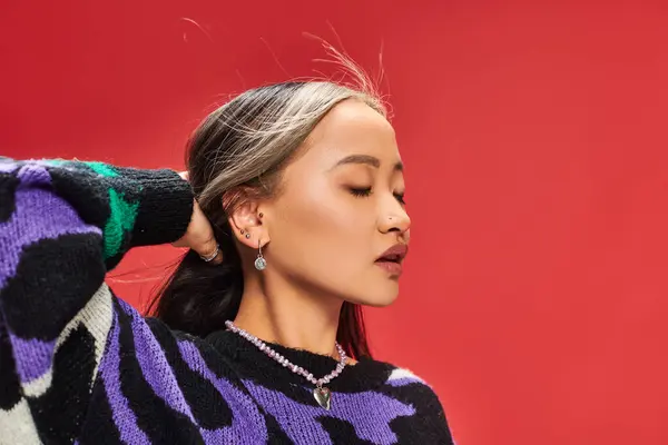Alluring young asian woman in vibrant sweater with animal print adjusting hair on red backdrop — Stock Photo