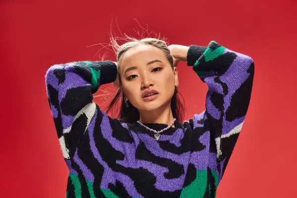 Beautiful young asian woman in vibrant sweater with animal print adjusting hair on red backdrop — Stock Photo