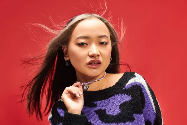 Pretty young asian woman in vibrant sweater with animal print touching necklace on red backdrop — Stock Photo