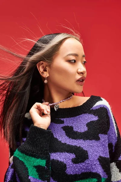 Charming young asian woman in vibrant sweater with animal print touching necklace on red backdrop — Stock Photo