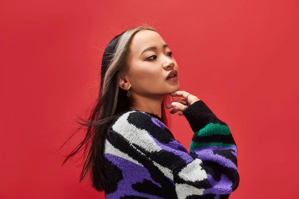 Beautiful young asian model in vibrant sweater with animal print touching chin on red backdrop — Stock Photo