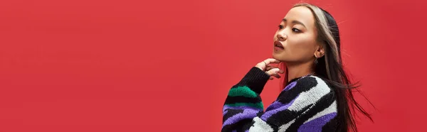 Schöne junge asiatische Modell in lebendigen Pullover mit Animal Print berühren Kinn auf rot, Banner — Stockfoto