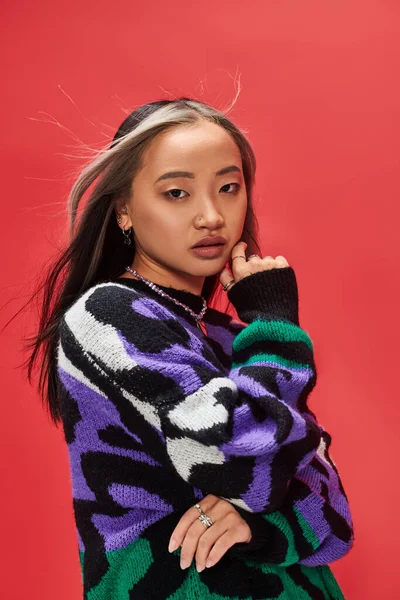 Beautiful young asian model in vibrant sweater with animal print touching cheek on red backdrop — Stock Photo