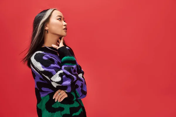 Schöne junge asiatische Modell in lebendigen Pullover mit Animal Print berühren Hals auf rotem Hintergrund — Stockfoto