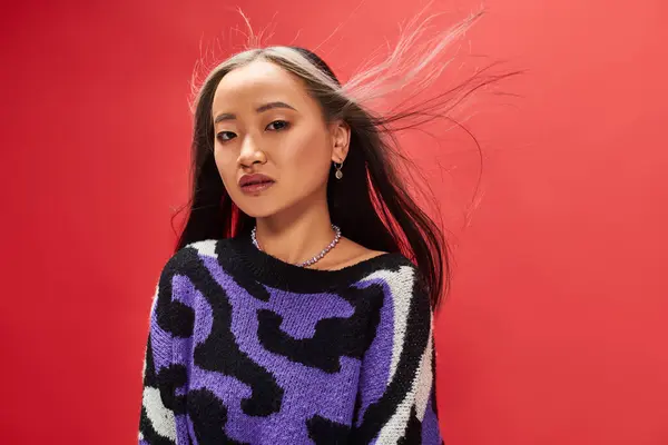 Wind in hair of beautiful young asian woman in sweater with animal print posing on red background — Stock Photo