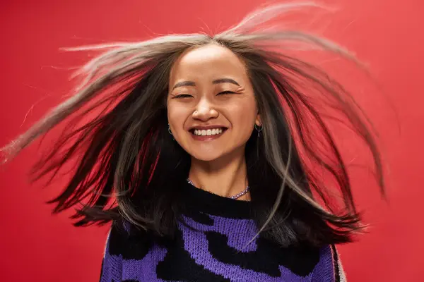 Wind in hair of joyful young asian woman in sweater with animal print posing on red background — Stock Photo