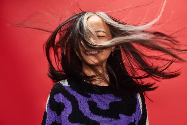 Вітер у волоссі задоволеної молодої азіатки в светрі з принтом тварин позує на червоному тлі — стокове фото