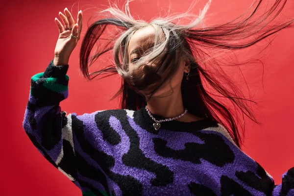 Wind in hair of young asian model in sweater with animal print gesturing on red background — Stock Photo