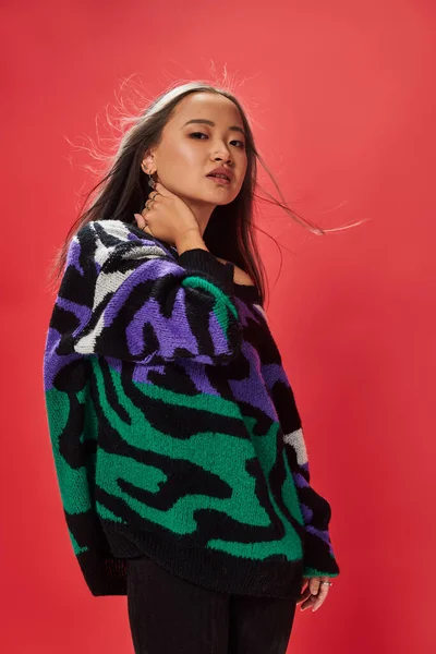 Young asian woman in sweater with animal print touching neck while posing on red backdrop — Stock Photo