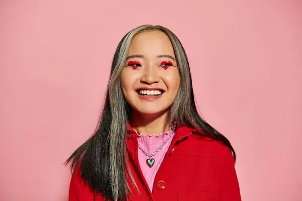 Valentines day concept, cheerful asian woman with heart shaped eye makeup posing on pink backdrop — Stock Photo