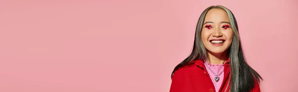 Banner de San Valentín, mujer asiática feliz con maquillaje de ojos en forma de corazón posando sobre fondo rosa - foto de stock