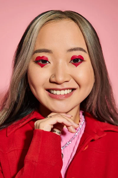 San Valentín concepto de día, feliz mujer asiática con el corazón en forma de maquillaje de ojos sonriendo en rosa telón de fondo - foto de stock
