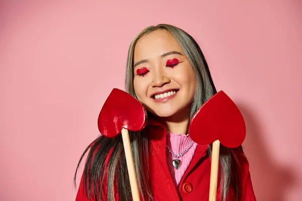 Valentines day concept, jolly asian girl with heart shaped eye makeup holding arrows on pink — Stock Photo