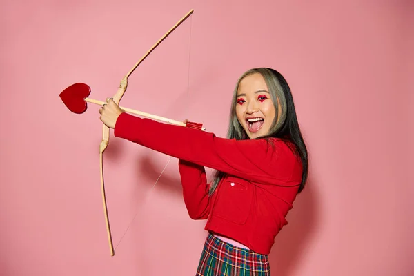 Valentines day, jolly asian woman with heart shaped eye makeup holding cupid arrow and bow on pink — Stock Photo