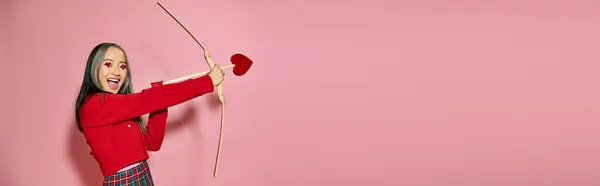 Saint Valentin, heureuse femme asiatique avec maquillage des yeux en forme de coeur tenant la flèche et l'arc de Cupidon sur rose — Photo de stock