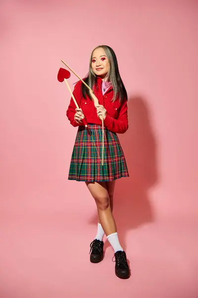 Día de San Valentín, alegre mujer asiática con el corazón en forma de maquillaje de ojos la celebración de la flecha de Cupido y arco en rosa - foto de stock