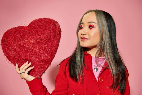 San Valentín día, alegre asiático mujer con vibrante ojo maquillaje celebración rojo corazón en rosa telón de fondo - foto de stock