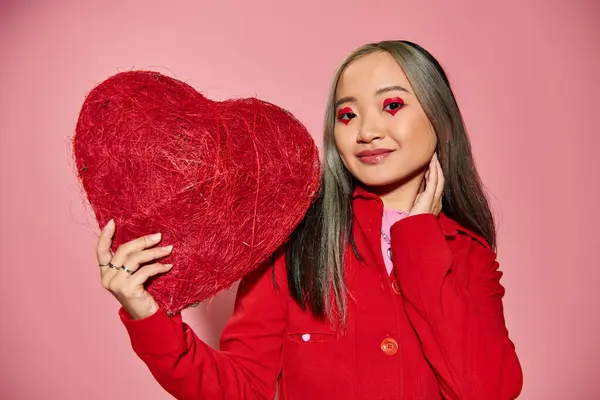 Valentine dia, feliz asiático mulher com vibrante olho maquiagem segurando vermelho coração em rosa pano de fundo — Fotografia de Stock