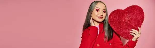 Día de San Valentín, mujer asiática feliz con el maquillaje vibrante del ojo sosteniendo el corazón rojo en el telón de fondo rosa, bandera - foto de stock