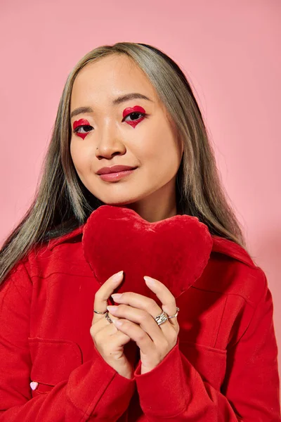 Valentine dia, alegre asiático mulher com vibrante olho maquiagem segurando vermelho coração no rosa pano de fundo — Fotografia de Stock