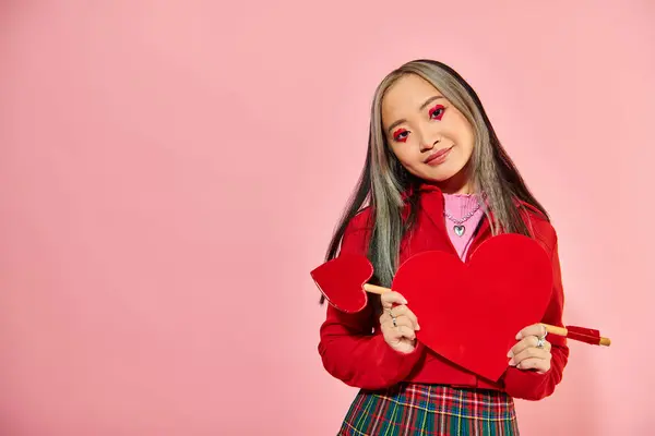 San Valentín día, sonriente mujer asiática con vibrante ojo maquillaje celebración de cartón corazón en rosa telón de fondo - foto de stock