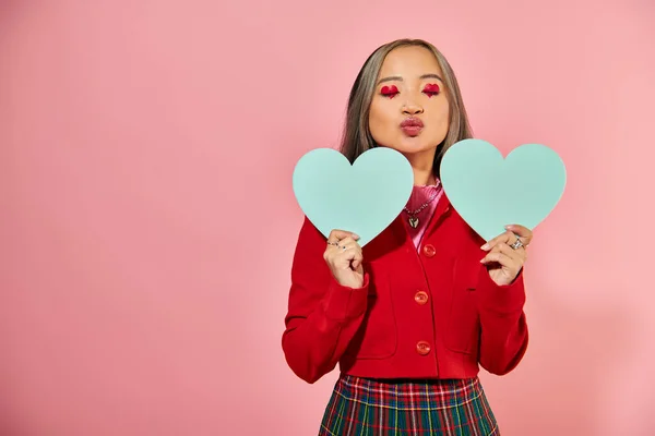 San Valentín día, joven asiático chica con vibrante ojo maquillaje celebración azul carton corazones en rosa telón de fondo - foto de stock