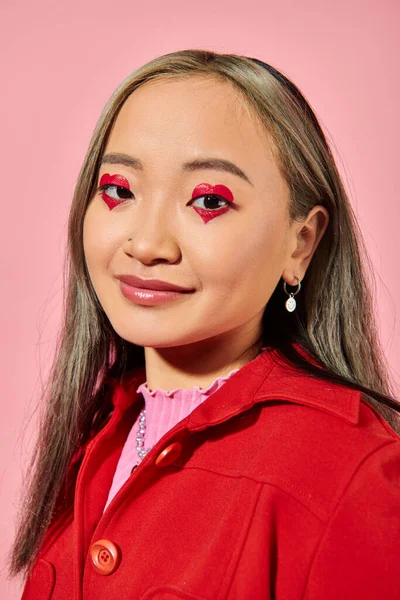 San Valentín día, sonriente asiática joven con el corazón ojo maquillaje posando en rojo chaqueta en rosa - foto de stock