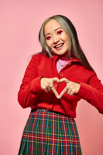 San Valentín concepto de día, feliz asiática chica en rojo chaqueta mostrando corazón con los dedos en rosa telón de fondo - foto de stock
