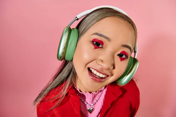 Happy young asian woman in green wireless headphones with heart shaped makeup posing on pink — Stock Photo