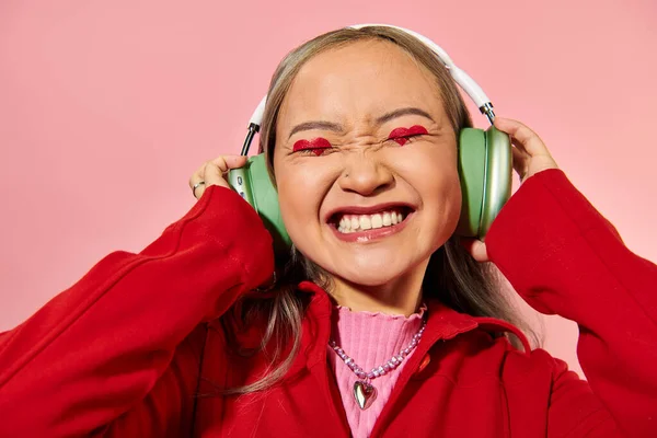 Alegre joven asiático mujer en verde inalámbrico auriculares con corazón en forma de maquillaje en rosa telón de fondo - foto de stock