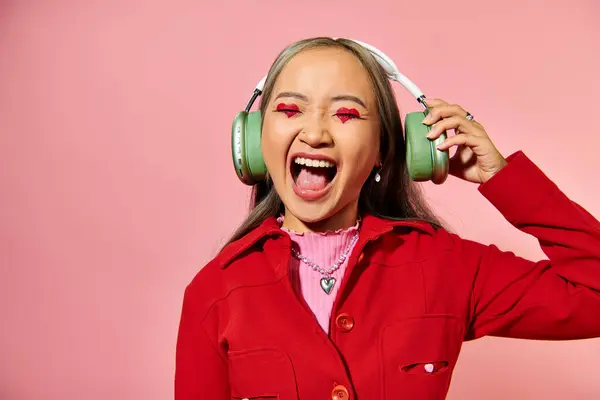 Excitada joven asiática mujer escuchando música en auriculares inalámbricos y gritando sobre fondo rosa - foto de stock