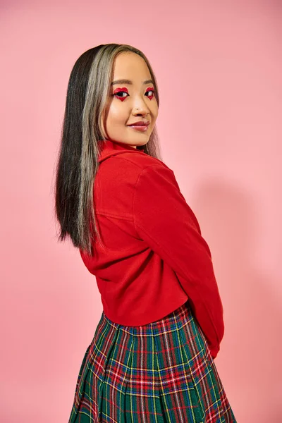 Asiático joven mujer en valentines día rojo chaqueta posando en rosa fondo, corazón forma ojo maquillaje - foto de stock