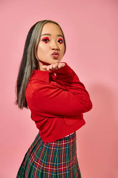 Asiático joven mujer en rojo chaqueta mostrando aire beso gesto en rosa fondo, corazón forma ojo maquillaje - foto de stock