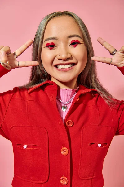 Happy asian young woman in red jacket showing v sign on pink background, heart shape eye makeup — Stock Photo