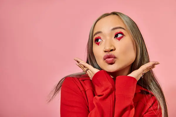 Bastante asiático joven mujer con corazón forma ojo maquillaje puting labios y mirando lejos en rosa telón de fondo - foto de stock