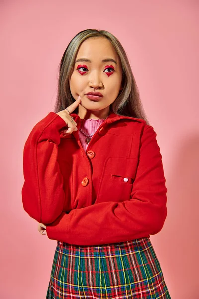Pensativo asiático joven mujer con corazón forma ojo maquillaje tocando mejilla y mirando lejos en rosa - foto de stock