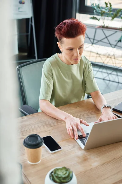 Стильна амбітна квір-людина, що працює в ноутбуці біля смартфона і кави, щоб піти в сучасному офісі — стокове фото