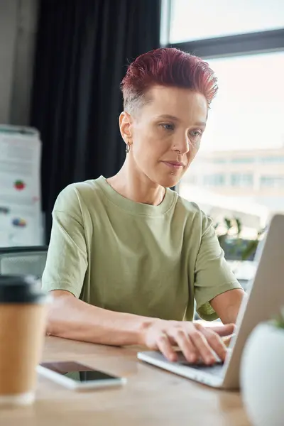 Stylish queer person working at laptop near blurred smartphone and coffee to go in modern office — Stock Photo