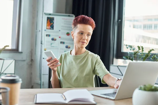 Elegante individuo bigender con smartphone che lavora al computer portatile vicino notebook in ufficio moderno — Foto stock