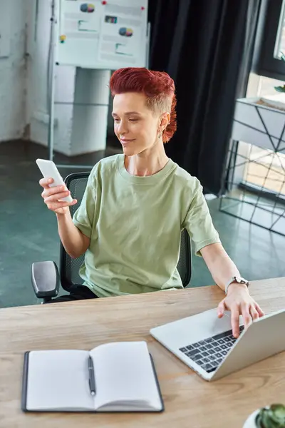 Smiling bigender individual looking at smartphone near laptop and notebook in modern office — Stock Photo