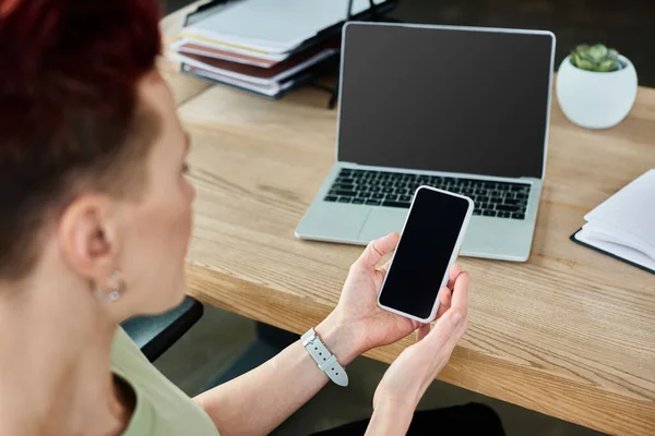 Persona d'affari non binaria che tiene smartphone con schermo vuoto vicino al computer portatile sul posto di lavoro in ufficio — Foto stock